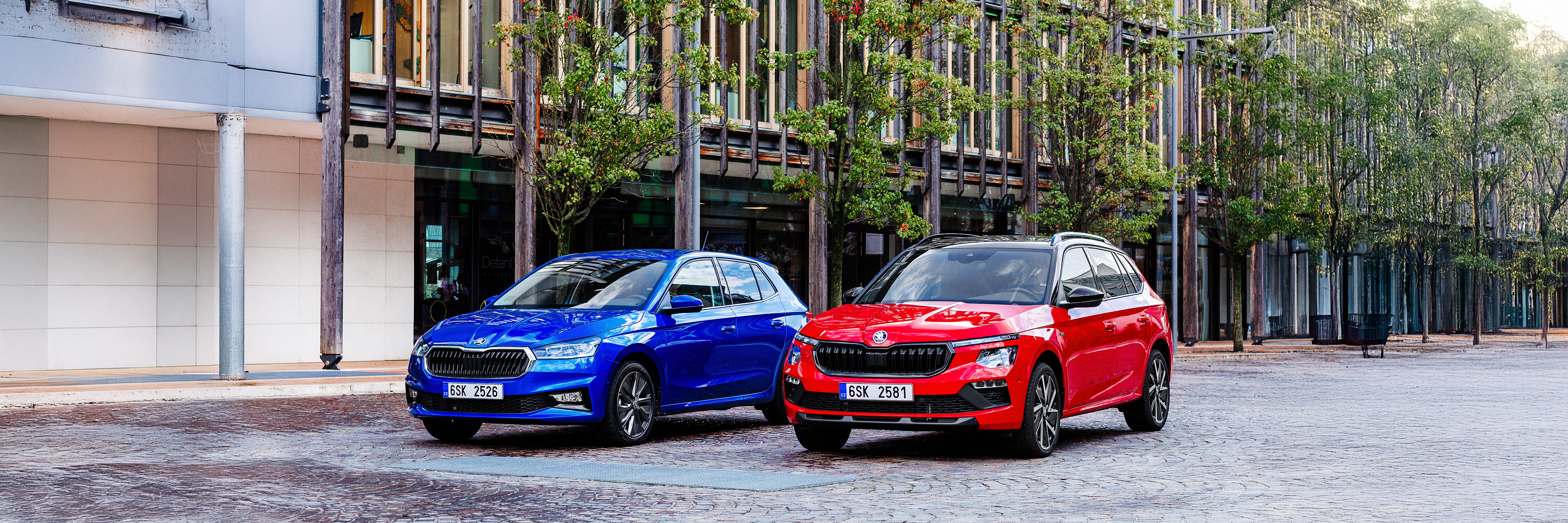 Na sliki sta Škoda Fabia in Škoda Kamiq, eden moder in eden rdeč, parkirana na tlakovani cesti pred moderno stavbo. Zraven stavbe rastejo drevesa.
