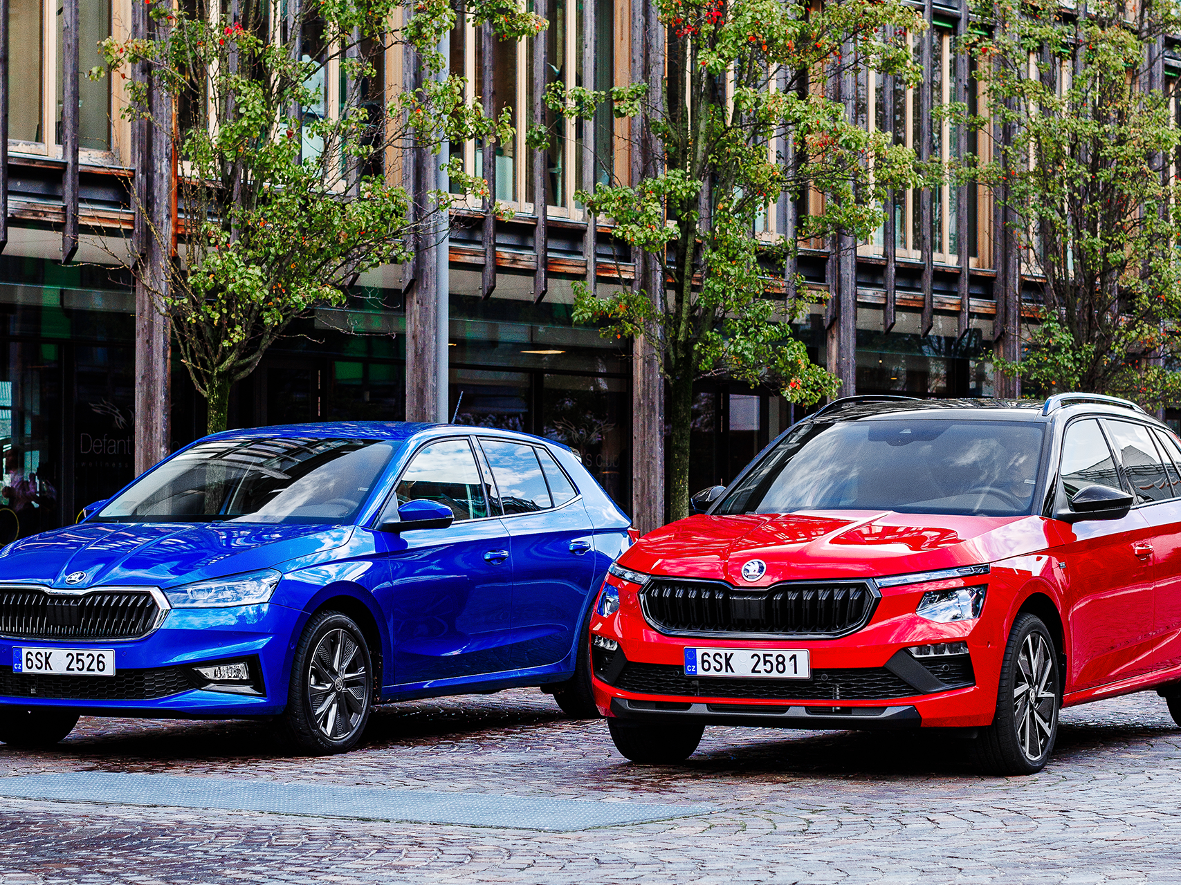 Na sliki sta Škoda Fabia in Škoda Kamiq, eden moder in eden rdeč, parkirana na tlakovani cesti pred moderno stavbo. Zraven stavbe rastejo drevesa.