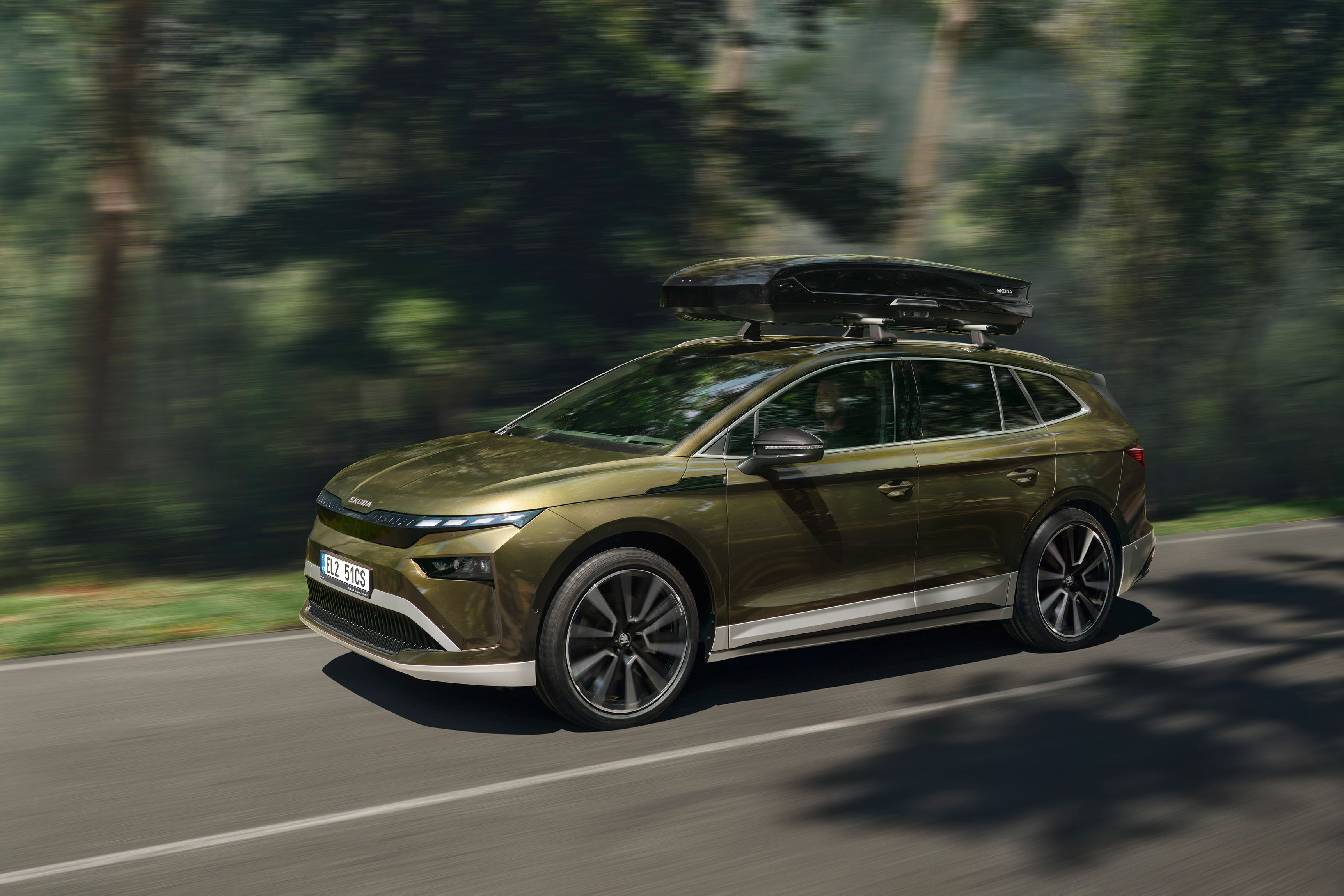 Avtomobil Kia EV9, ki vozi po cesti skozi gozdno območje. Vozilo je zeleno, aerodinamično oblikovano, na strehi ima pritrjeno strešno kovček. Cesta je ovita v bujno vegetacijo.