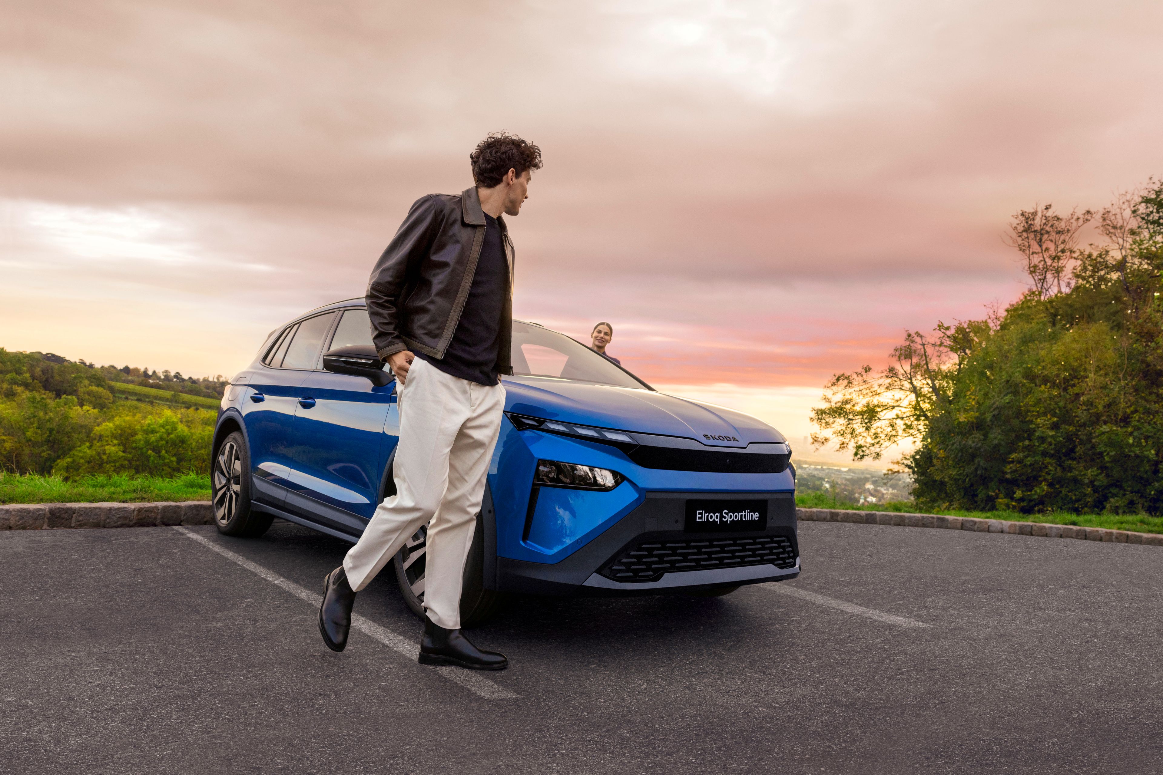 Moški stoji ob modrem električnem avtomobilu na parkirišču. V ozadju je gozdnata pokrajina in oblačno nebo med sončnim zahodom.