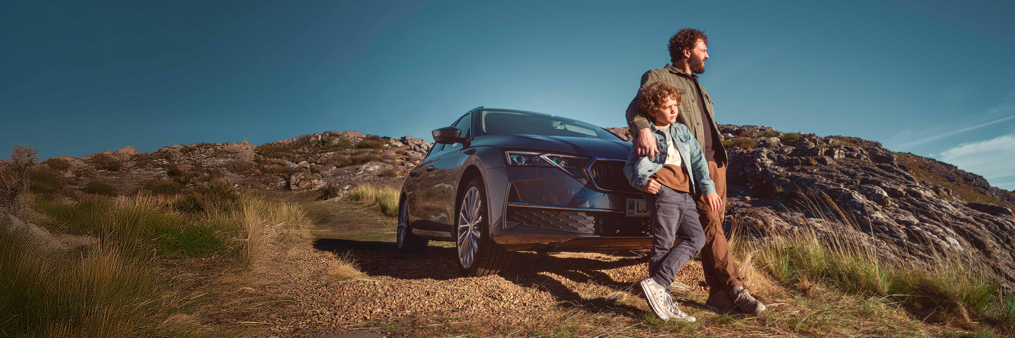 Oseba in otrok stojita na podeželski poti ob parkiranem avtomobilu Škoda Octavia Combi, obdanem z skalami in travo pod jasnim modrim nebom.