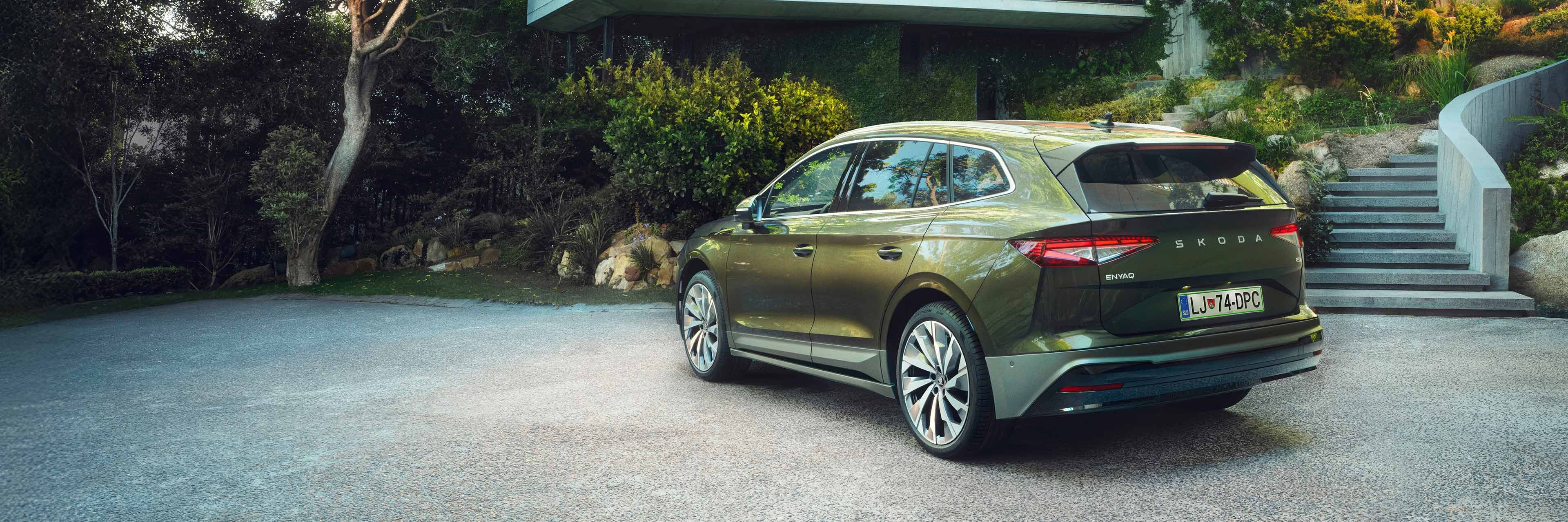 Zeleni Škoda Enyaq parkiran na dovozu pred moderno hišo, obkrožen z lepo urejeno zelenico in grmovjem.