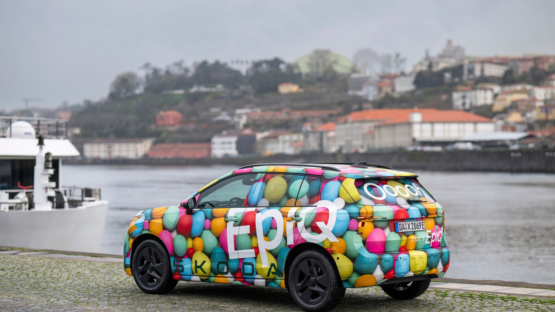 Vozilo Škoda Epiq, parkirano ob reki, ima živahno poslikavo s pisanimi baloni in besedami kot so "Epic" in "Škoda". V ozadju je vidno mesto s starimi zgradbami. Ob reki stoji tudi ladja.