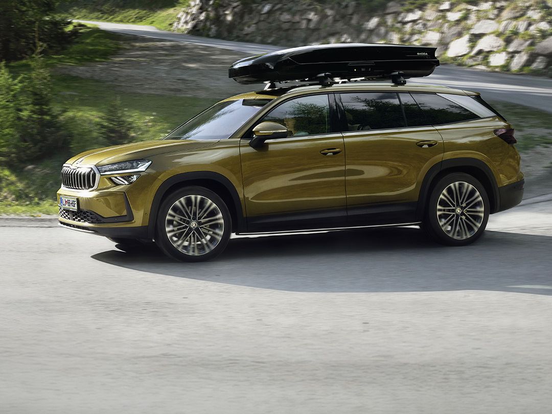 Rjav SUV Škoda Kodiaq z nameščenim strešnim kovčkom vozi po ovinkasti cesti sredi gozda. V ozadju so vidna drevesa in griči.