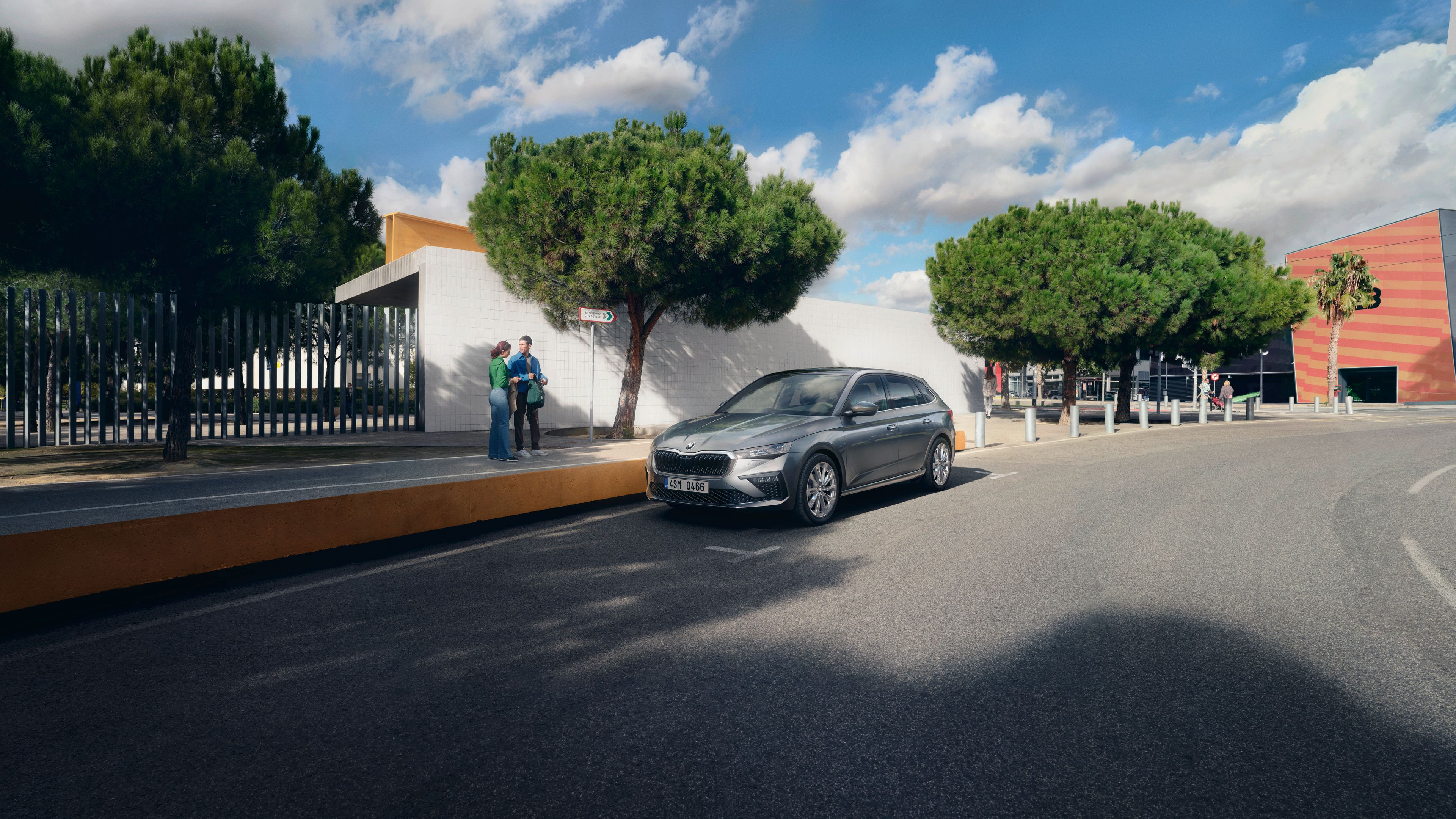 Siva limuzina je parkirana ob drevoredu na sončen dan. V ozadju sta dve osebi blizu vhoda v stavbo. Nebo je modro s puhastimi oblaki.