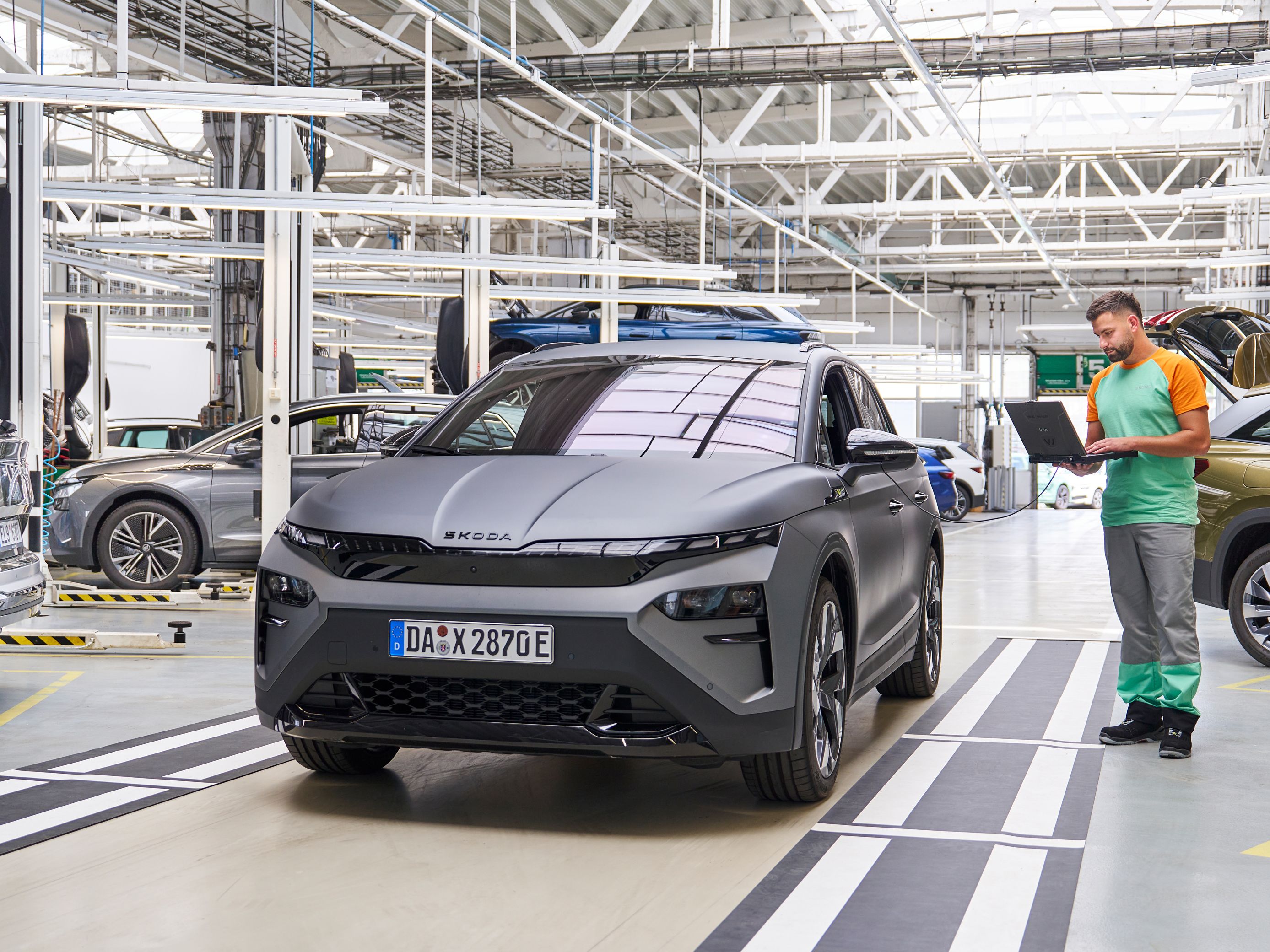 Siva avtomobil stoji v delavnici, medtem ko oseba v delovni uniformi zapiše nekaj v beležko. V ozadju so še drugi avtomobili na dvigalih, ki se očitno servisirajo.