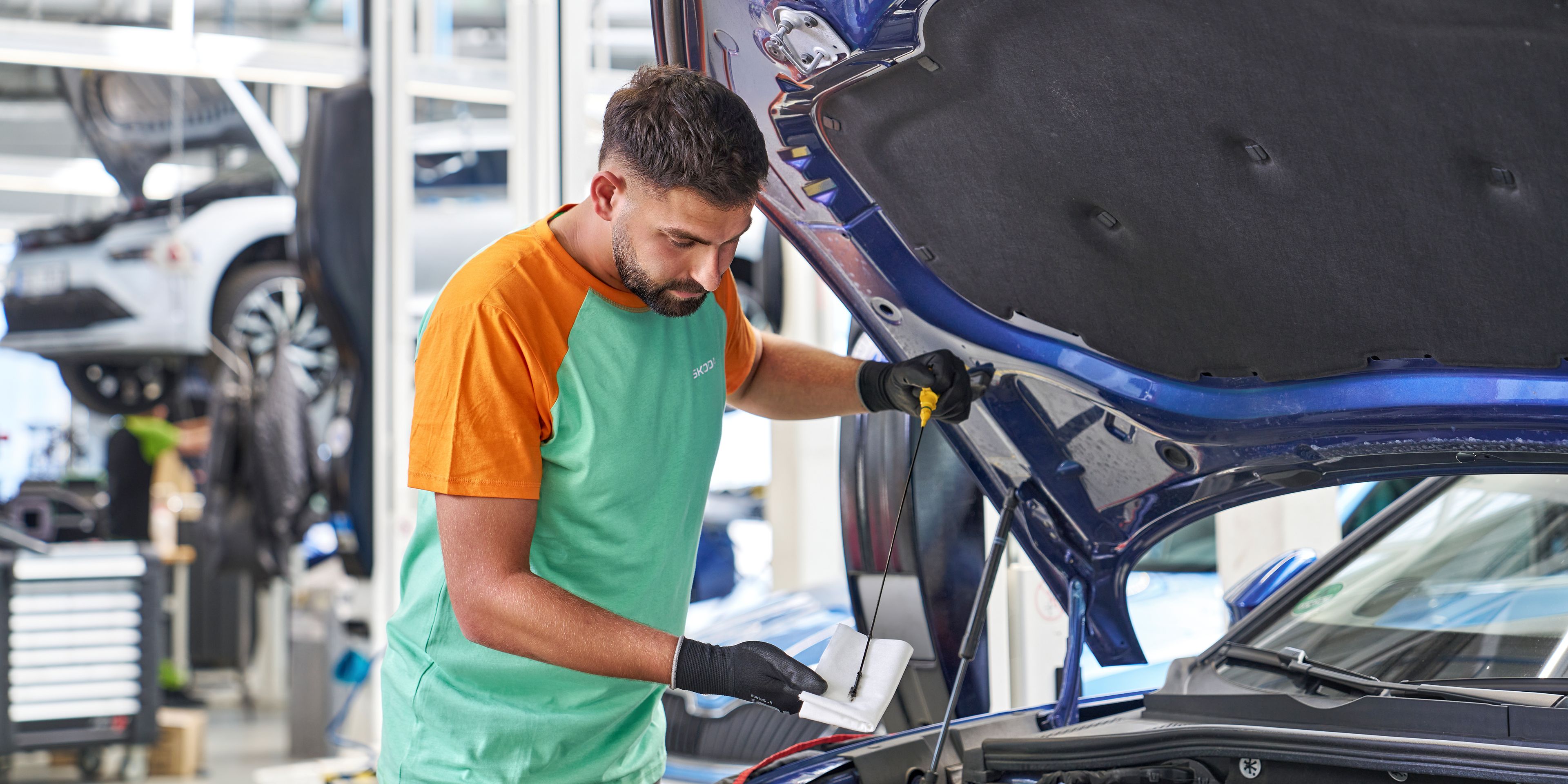 Mehanik pregleduje motor avtomobila v servisni delavnici. Drži list papirja in pritiska na motor. V ozadju so vidni drugi avtomobili in delovno orodje.