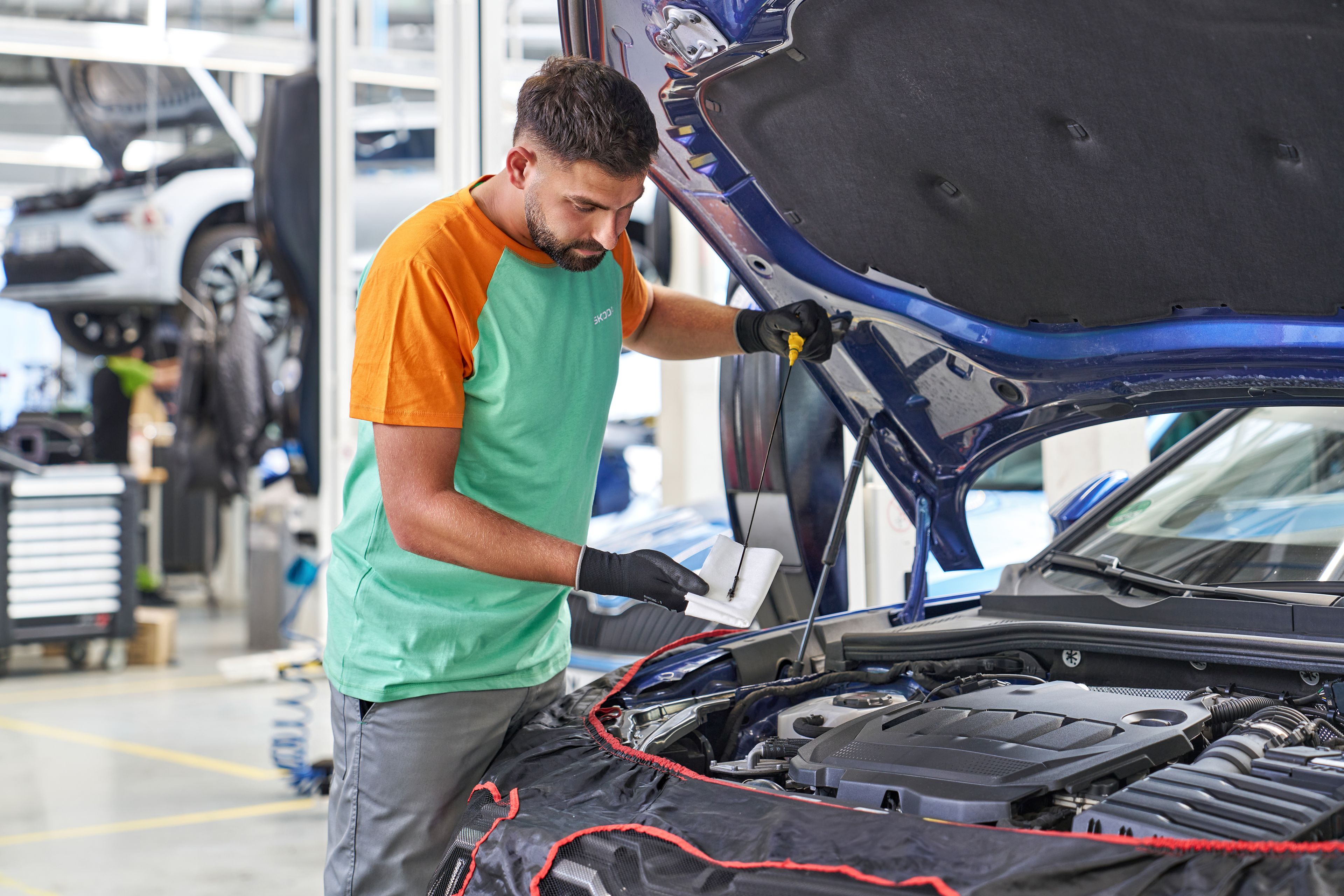Mehanik pregleduje motor avtomobila v servisni delavnici. Drži list papirja in pritiska na motor. V ozadju so vidni drugi avtomobili in delovno orodje.
