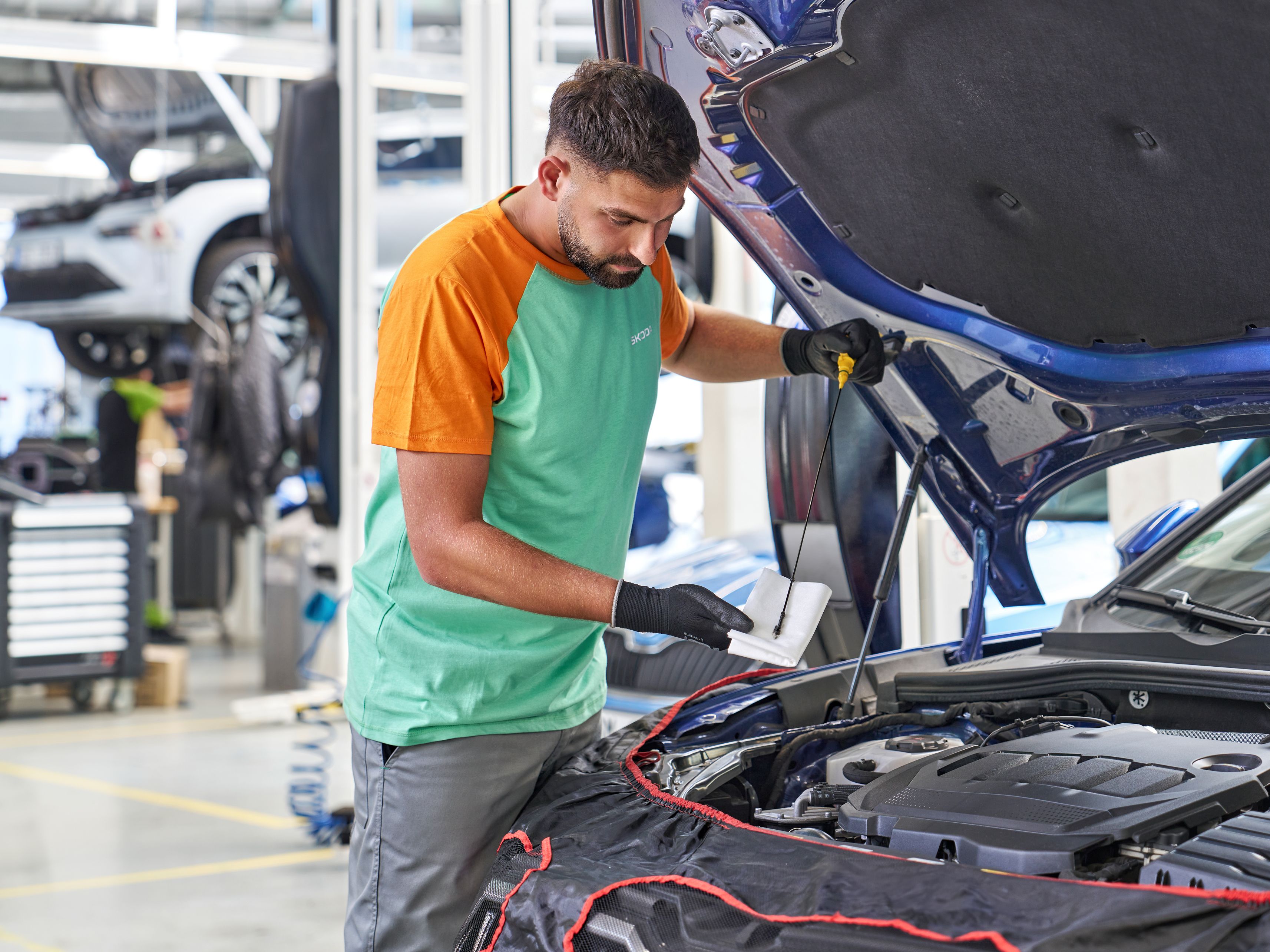 Mehanik pregleduje motor avtomobila v servisni delavnici. Drži list papirja in pritiska na motor. V ozadju so vidni drugi avtomobili in delovno orodje.