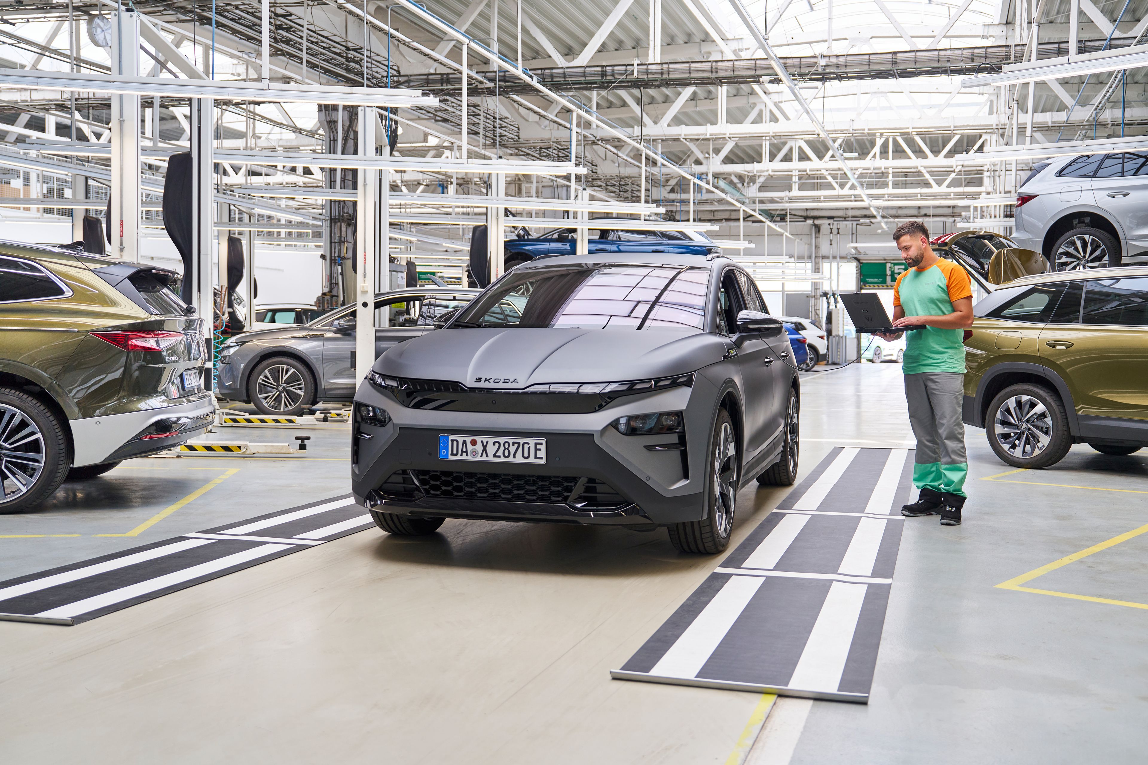 Siva avtomobil stoji v delavnici, medtem ko oseba v delovni uniformi zapiše nekaj v beležko. V ozadju so še drugi avtomobili na dvigalih, ki se očitno servisirajo.