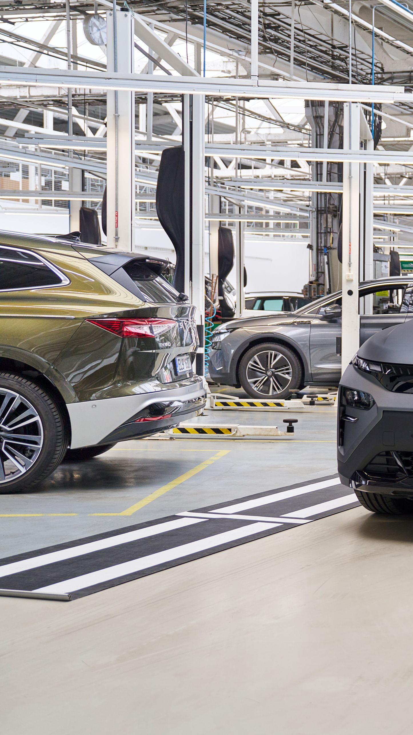 Siva avtomobil stoji v delavnici, medtem ko oseba v delovni uniformi zapiše nekaj v beležko. V ozadju so še drugi avtomobili na dvigalih, ki se očitno servisirajo.