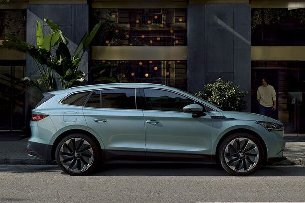 Škoda Enyaq parkiran ob ulici pred moderno stavbo. Ljudje se sprehajajo po pločniku.