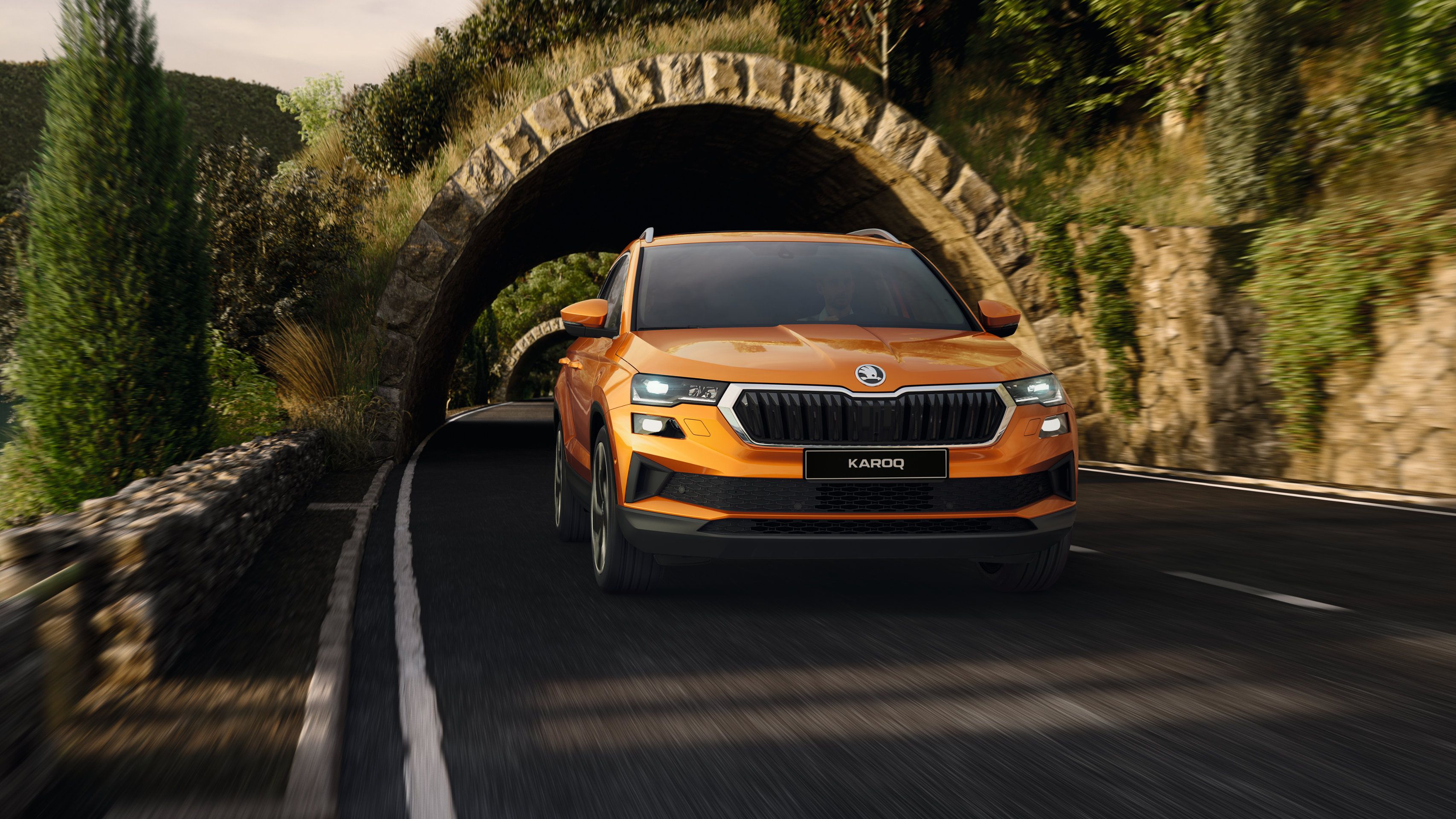 Oranžni Škoda Karoq prečka kamnit tunel na slikoviti gorski cesti. Avto izstopa s sodobnim dizajnom in aktivnimi lučmi. Ozadje prikazuje zeleno naravo in strme pečine.