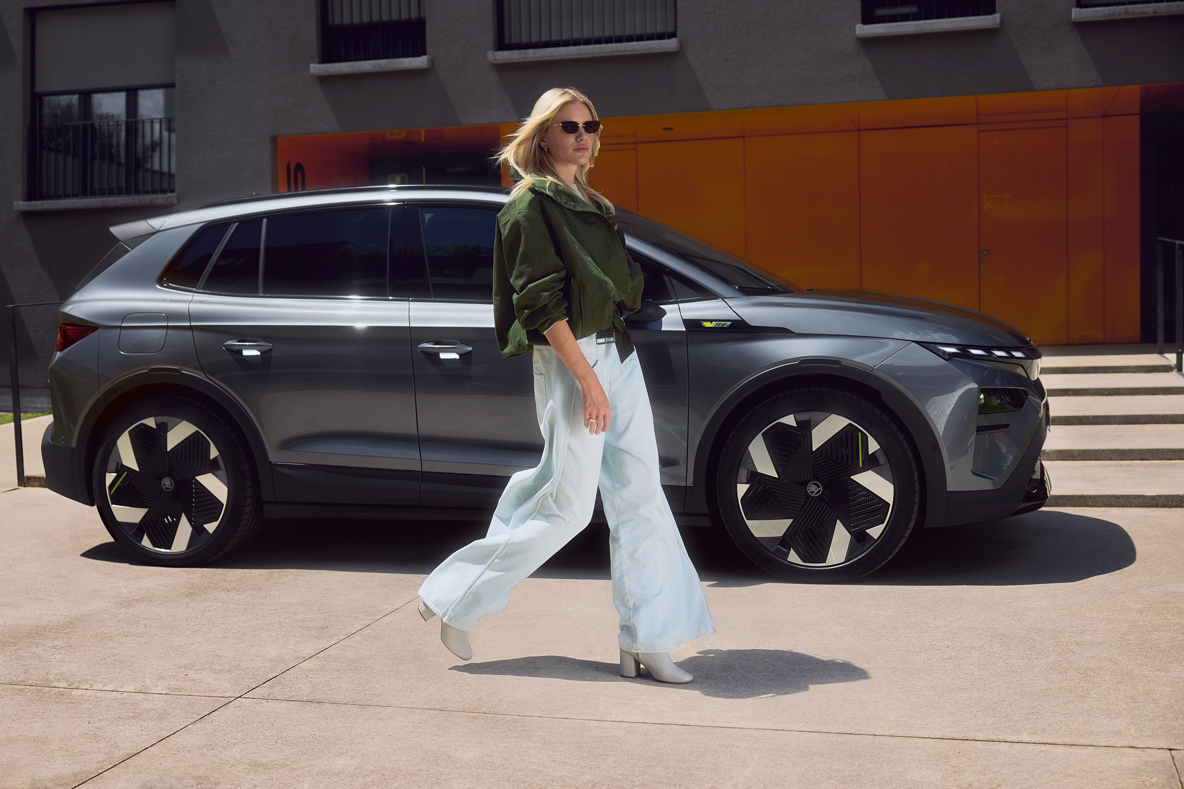 Oseba z zamegljenim obrazom hodi mimo moderne sivega športnega terenskega vozila. Oseba nosi široke bele hlače in zeleno jakno. Avto stoji pred zgradbo s sivimi stenami in oranžnimi vrati.